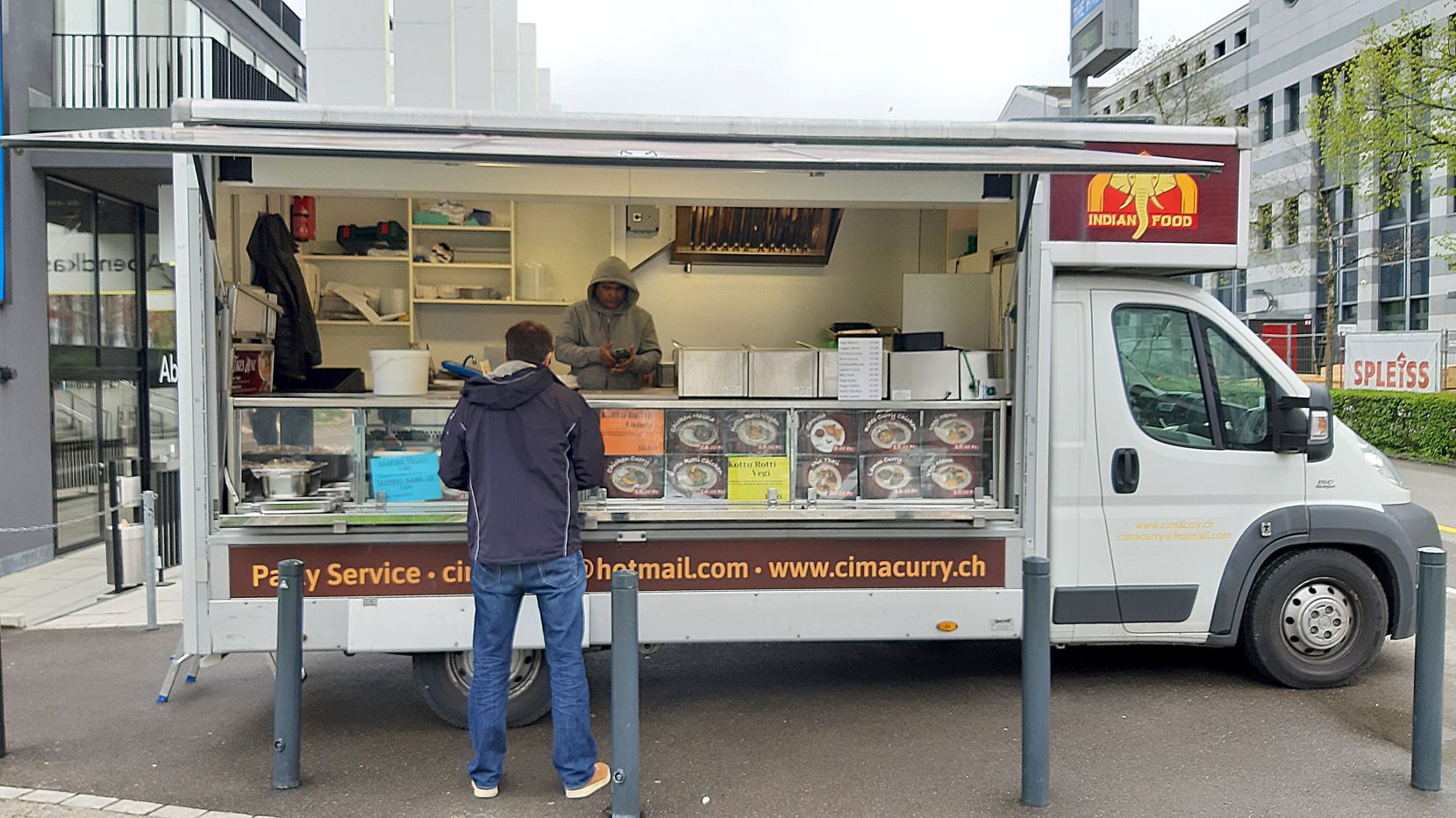 Ein Curry-Foodtruck in einem Gewerbegebiet, davor wartet ein Gast.